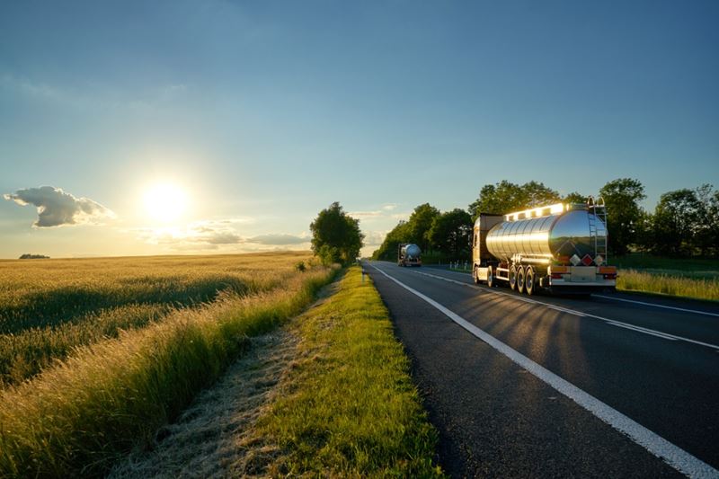 Trucks with chrome tanks driving on an asphalt road along the corn field at sunset.